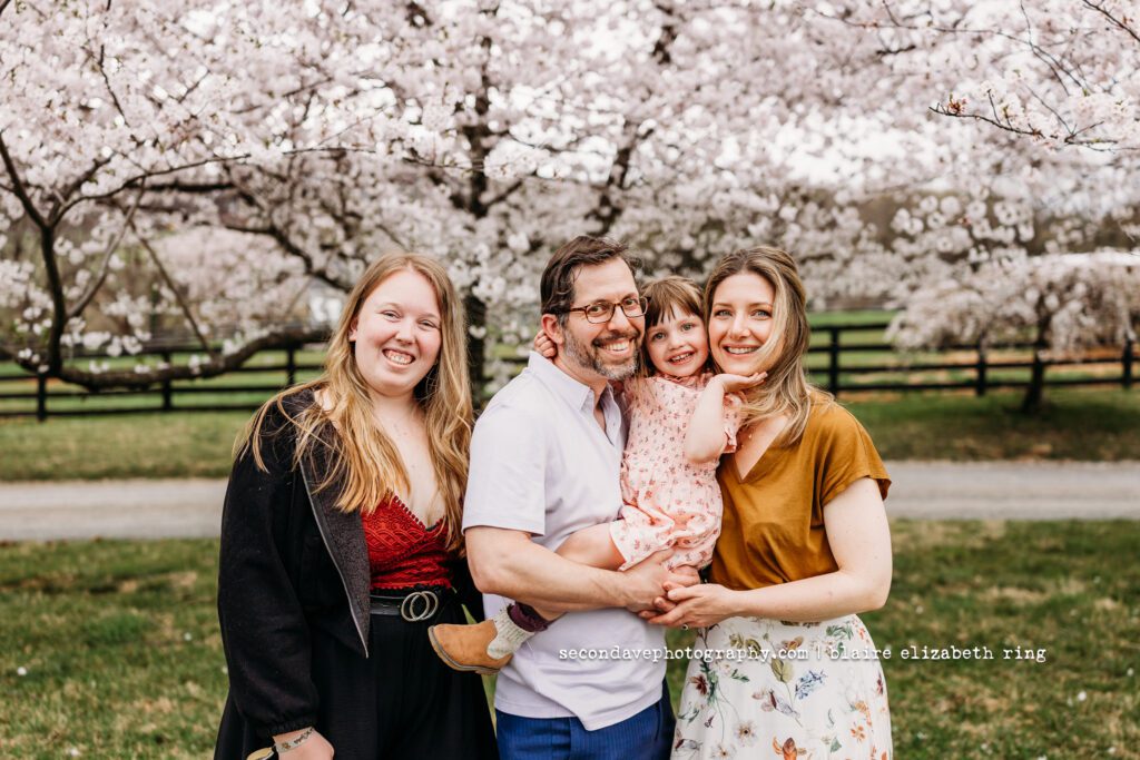Family of 3 and au pair smiling under blooming cherry blossom trees in Loudoun County.
