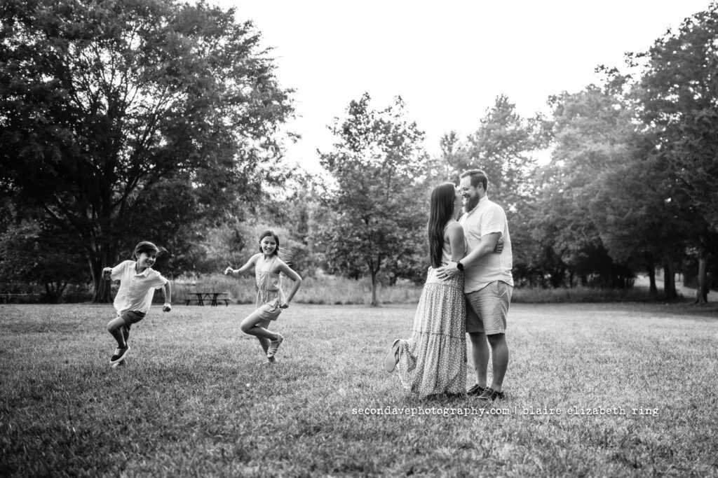 parents hugging and kids running circles around them photographed in black and white in sterling va