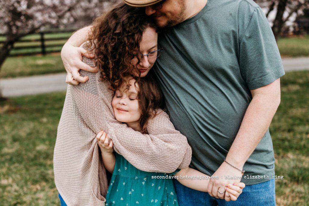Family of 3 in front of blooming cherry blossoms in Northern Virginia.
