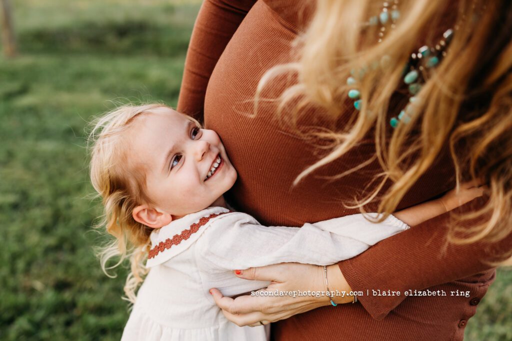 Toddler hugging pregnant mom's belly and looking up and smiling.